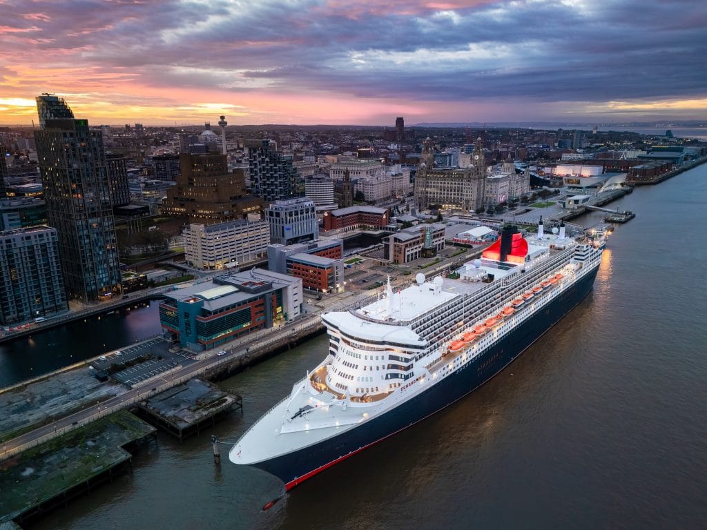 Aerial view of the Cunard cruise liner Queen Mary 2 docked in Liverpool at sunrise, with the city skyline in the background.