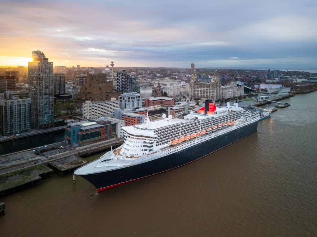 Aerial view of the Cunard cruise liner Queen Mary 2 docked in Liverpool, England, with the city skyline in the background at sunrise.
